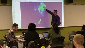 person pointing to image on projector screen while participants sit around tables