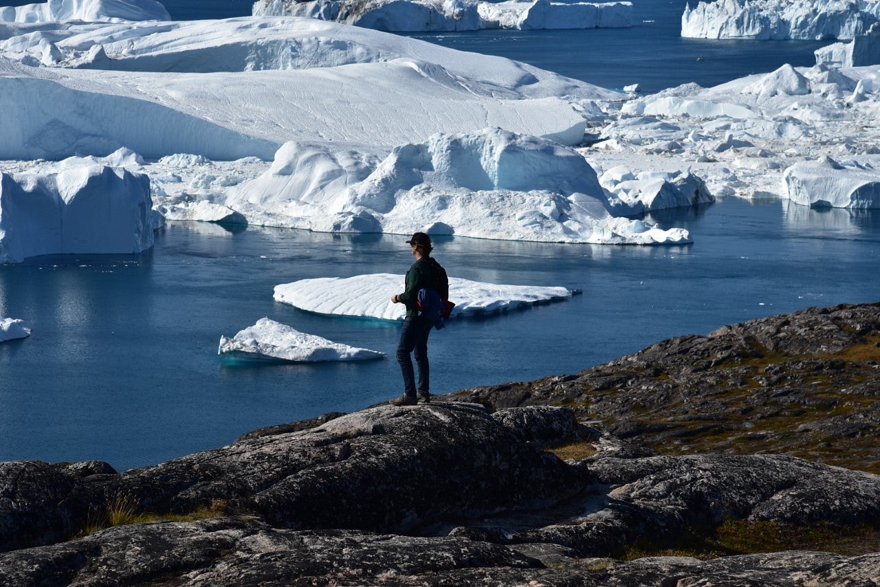 Man stands on edge of mountain looking at icebergs. 