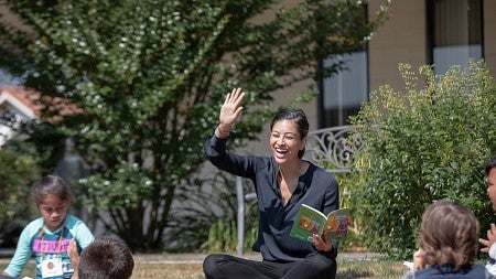 Teacher raises hand with children sitting in circle