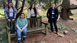 Students sit and stand around a bench outdoors 