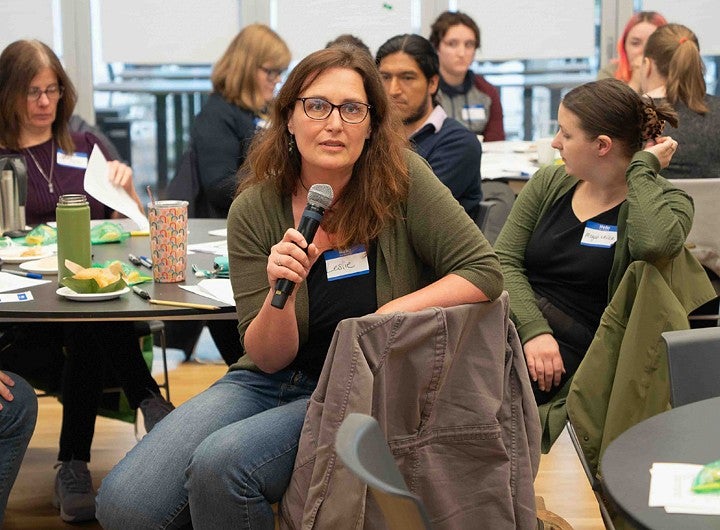 A person wearing glasses speaks into a microphone while seated at a table