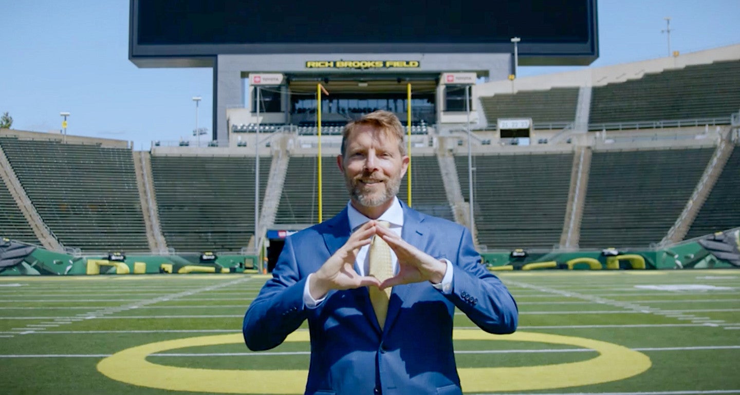 a person does the O hand gesture with the big screen mighty oregon behind him