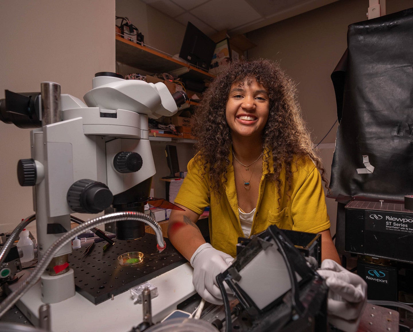 a person beside a microscope who is smiling with long curly hair 