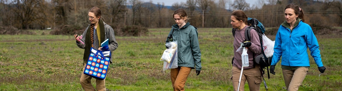 four women walking westward in a grassy area collecting soil