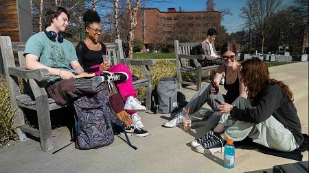 four students gathered, two on a bench, two sitting on the sidewalk