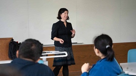 woman standing and teaching in front of a room of students sitting at tables