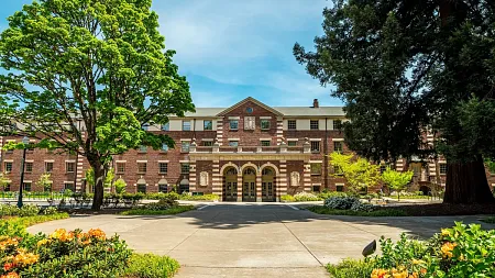 a brick facade building stands between lush green trees and blue skies