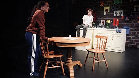 woman standing at table and man sitting on counter in a production for UO theatre arts