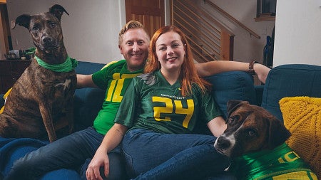 psychology alum mara sargent and her partners dressed in university of oregon gear sit on a couch with their dogs