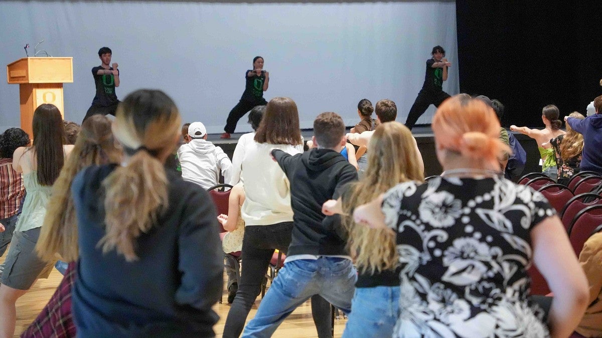 students practicing Wushu, watching demonstrators in the background on a stage