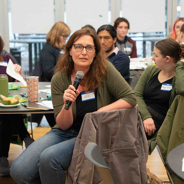 a woman wearing glasses speaks into a microphone while seated at a table